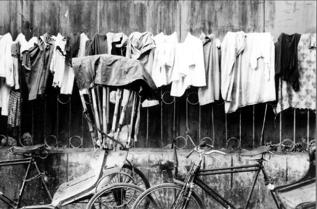 A black and white photo of a manual hand pulled rickshaw in Kolkata, India, parked in front of some laundry on a gate