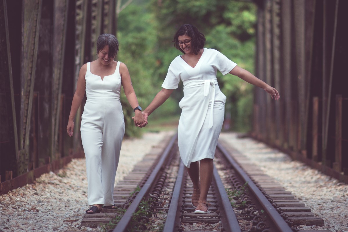 a color photo of a queer female couple in wedding whites walking along an old railway bridge