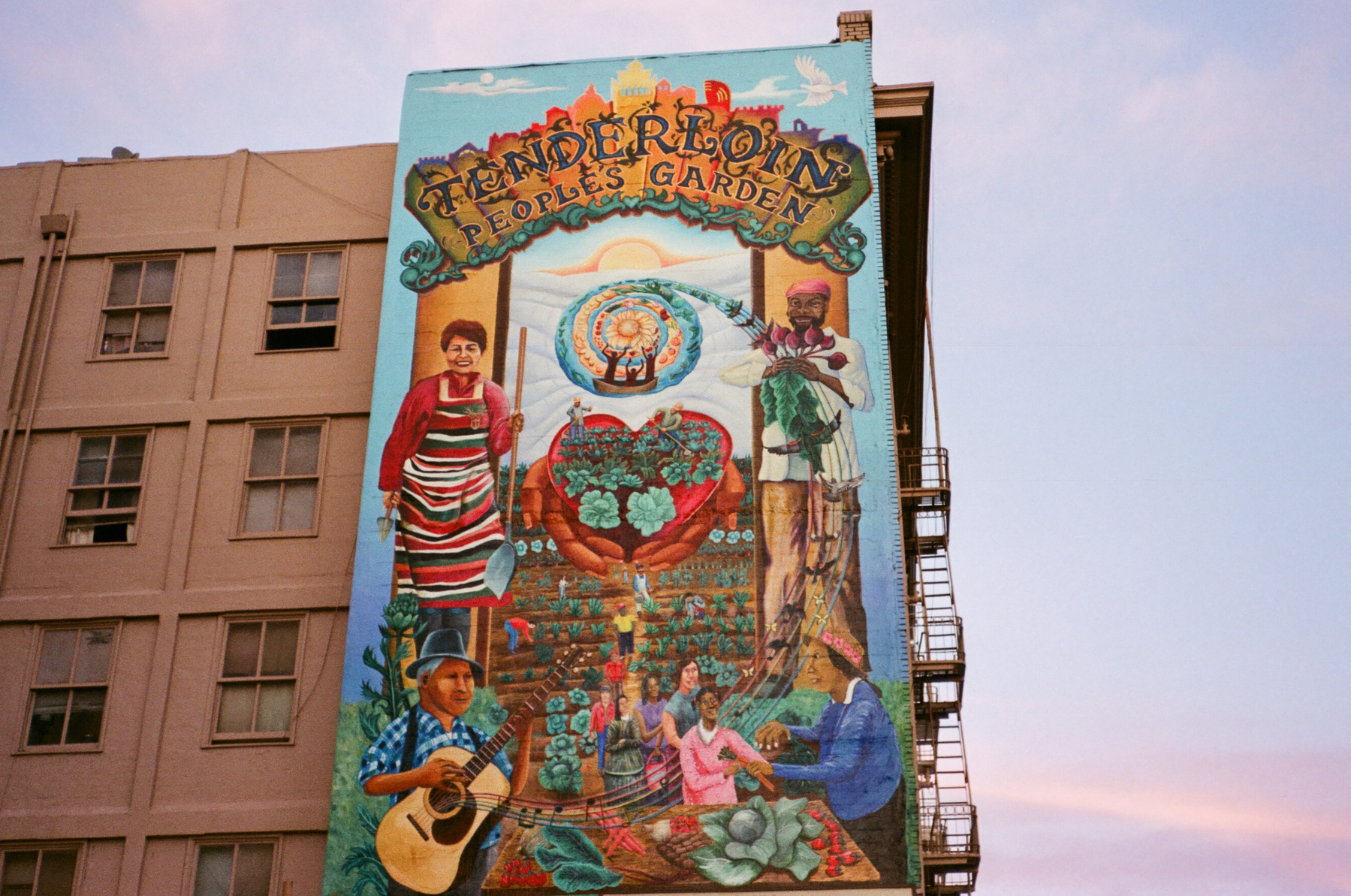A color photograph of a large mural which reads Tenderloin People's Garden on a building. Blue and pink sky in the background