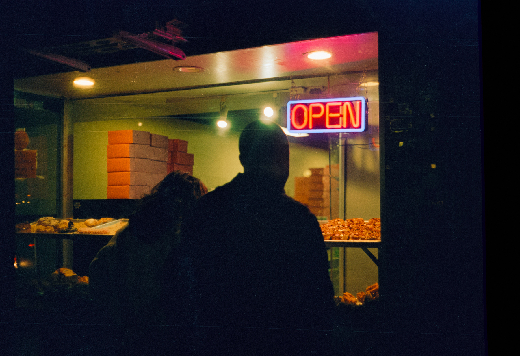 a scan of a color photograph that shows a night time scene of a couple looking through the windows of a donut shop