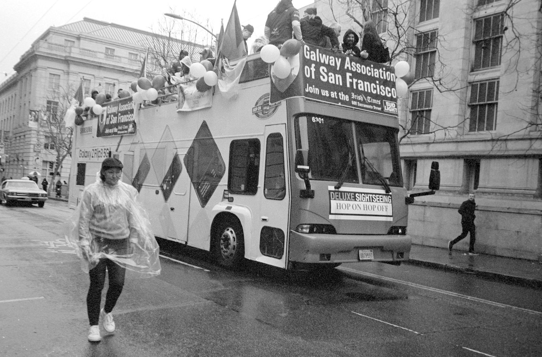a scan of a black and white photograph of a bus that says Galway Association of San Francisco with people in it, and a person walking alongside the bus with a poncho
