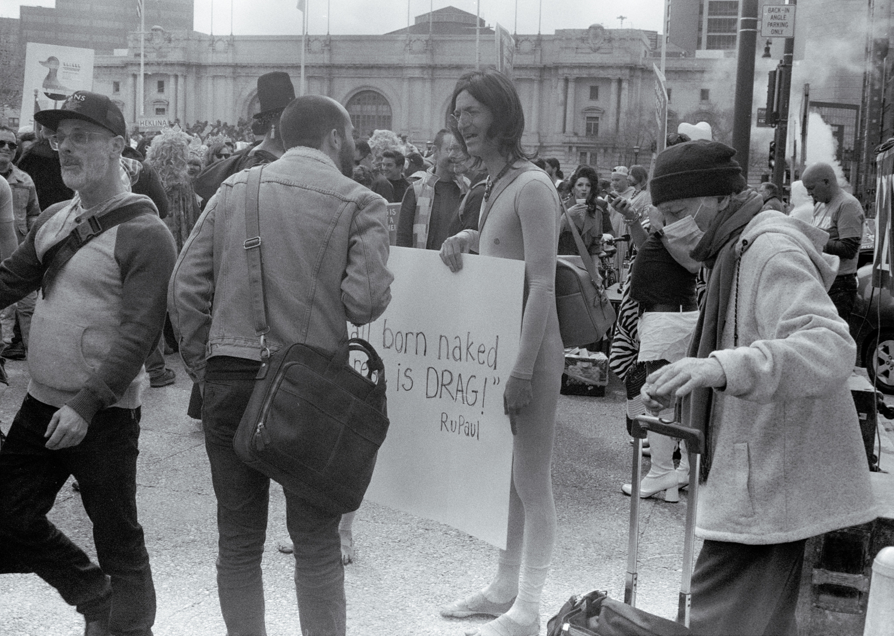 a scan of a black and white photo showing a person being interviewed, holding a sign that says Being Born Naked is a DRAG, a quote from Rupal