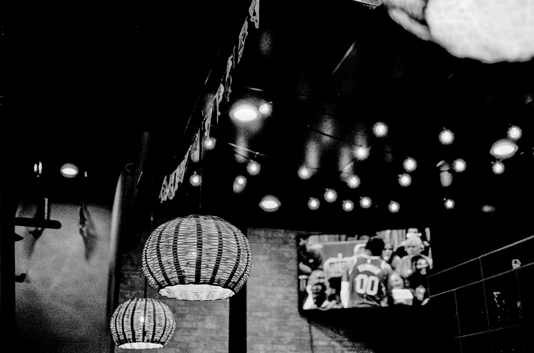 a scan of a black and white photo showing a few lamps at a restaurant with a woven pattern, in front of a TV showing NBA basketball