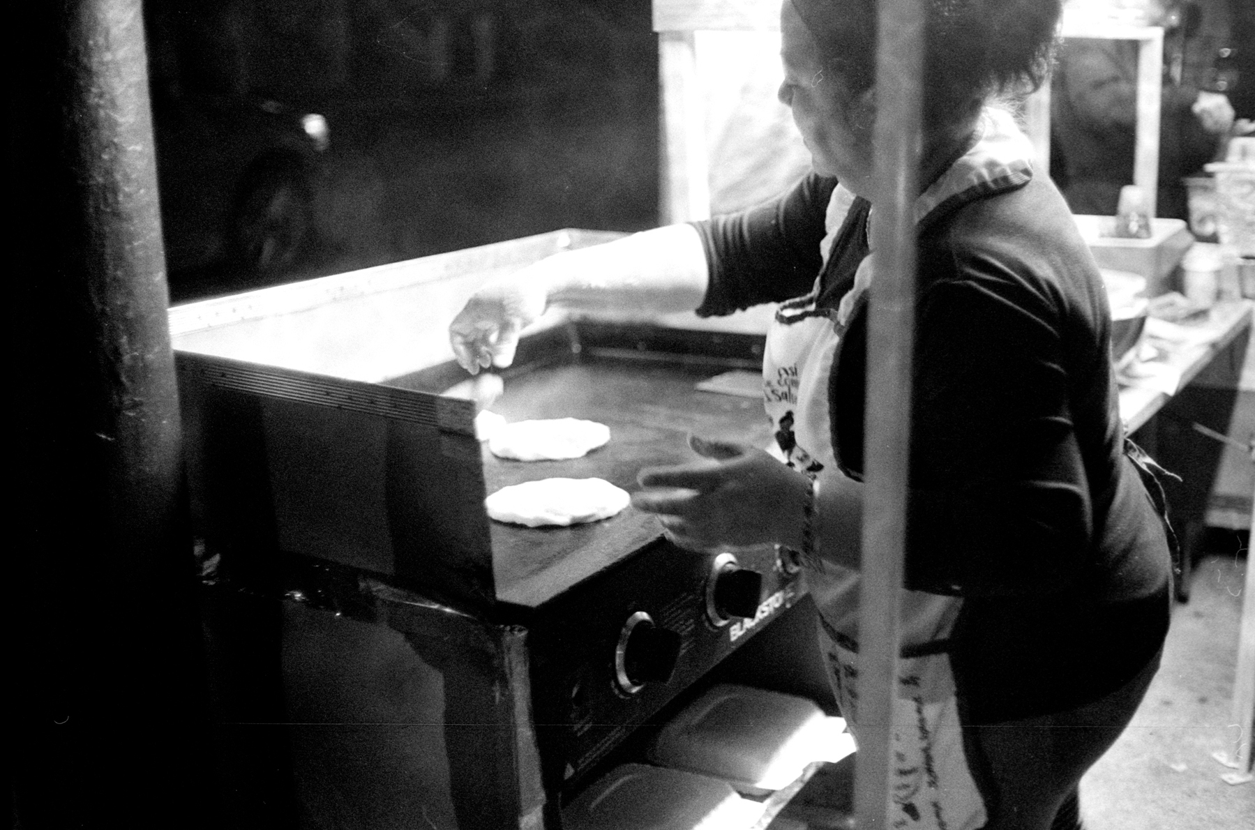 a scan of a black and white photo showing showing a lady cooking pupusa on a flat grill