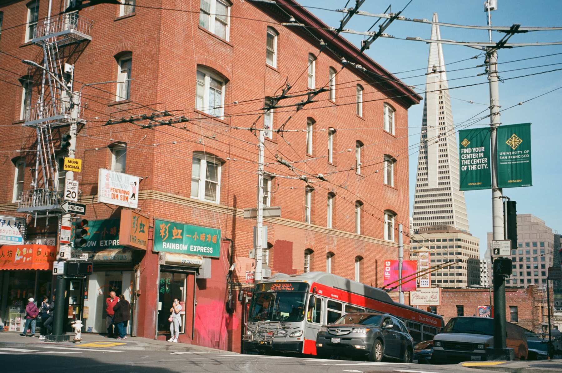 a color photo of the border of Chinatown San Francisco and the Financial District with Chinese shops in the foreground and TransAmerica pyramid in the background, a Muni bus at the bus stop at an intersection, people standing around or crossing the street