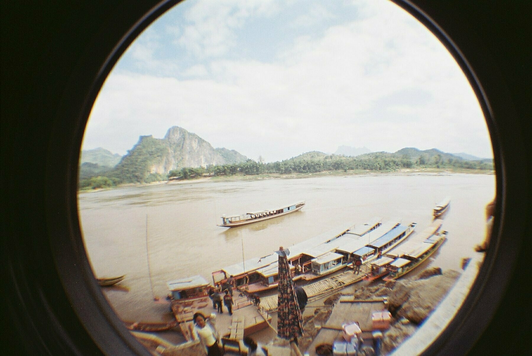a scan of a color photograph shot with a fish eye lens showing the river and boats outside a buddhist cave in Laos in Southeast Asia