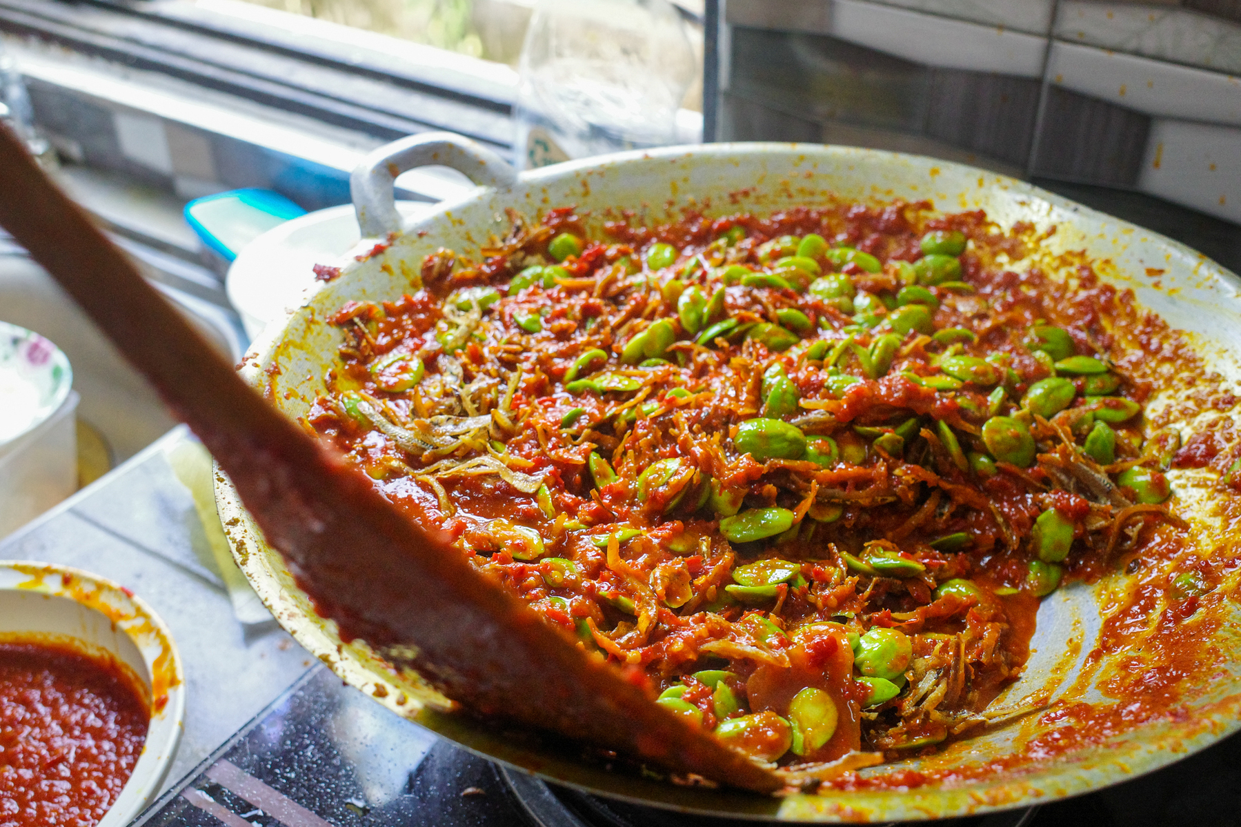 a digital photograph of a large wok full of chillies and stink beans being cooked into a sambal