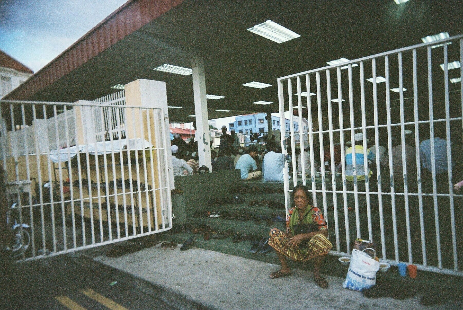a scan of a color photo of a woman sitting outside a mosque in singapore with lots of shoes behind her