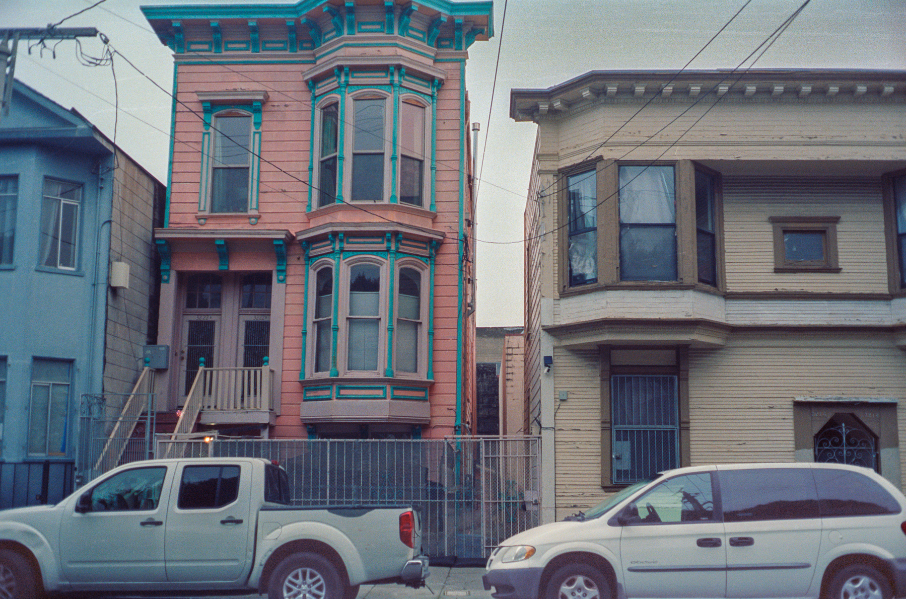 a scan of a color photograph of a pink and blue Victorian building on a street in San Francisco with two cars parked in front of it, and a yellow building next to it