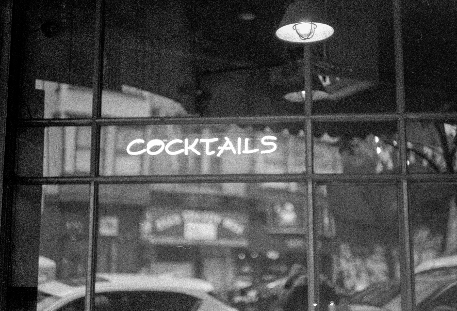 a scan of a black and white photo showing a neon sign that reads Cocktails in the window of a divey bar