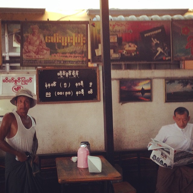 a photo of two people in a Burmese restaurant, one wearing a hat and another wearing traditional Burmese attire and reading a newspaper