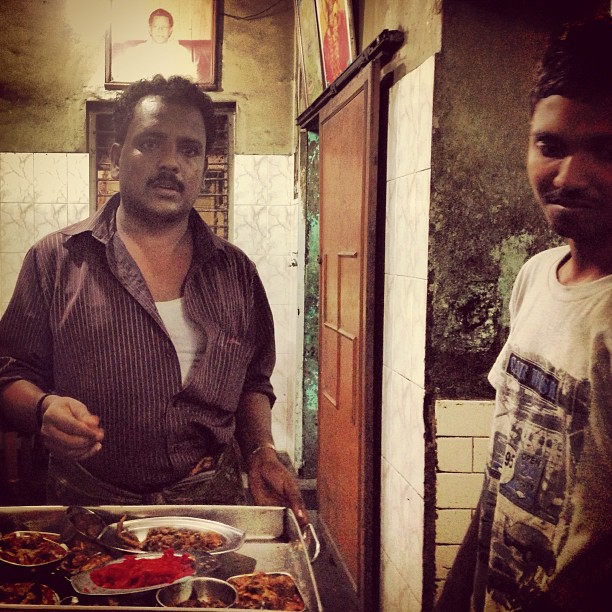 a photo of two men in Chennai, India, in a restaurant showing some food from behind a counter