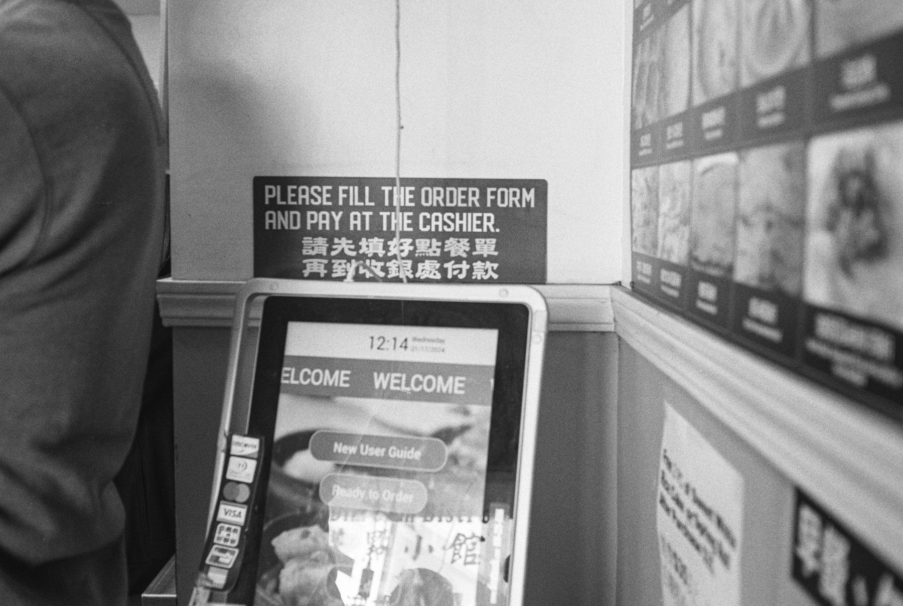 a scan of a black and white photo of a machine in a dimsum shop that takes orders and payment. on the wall, menu pictures and items and a person leaning on the wall