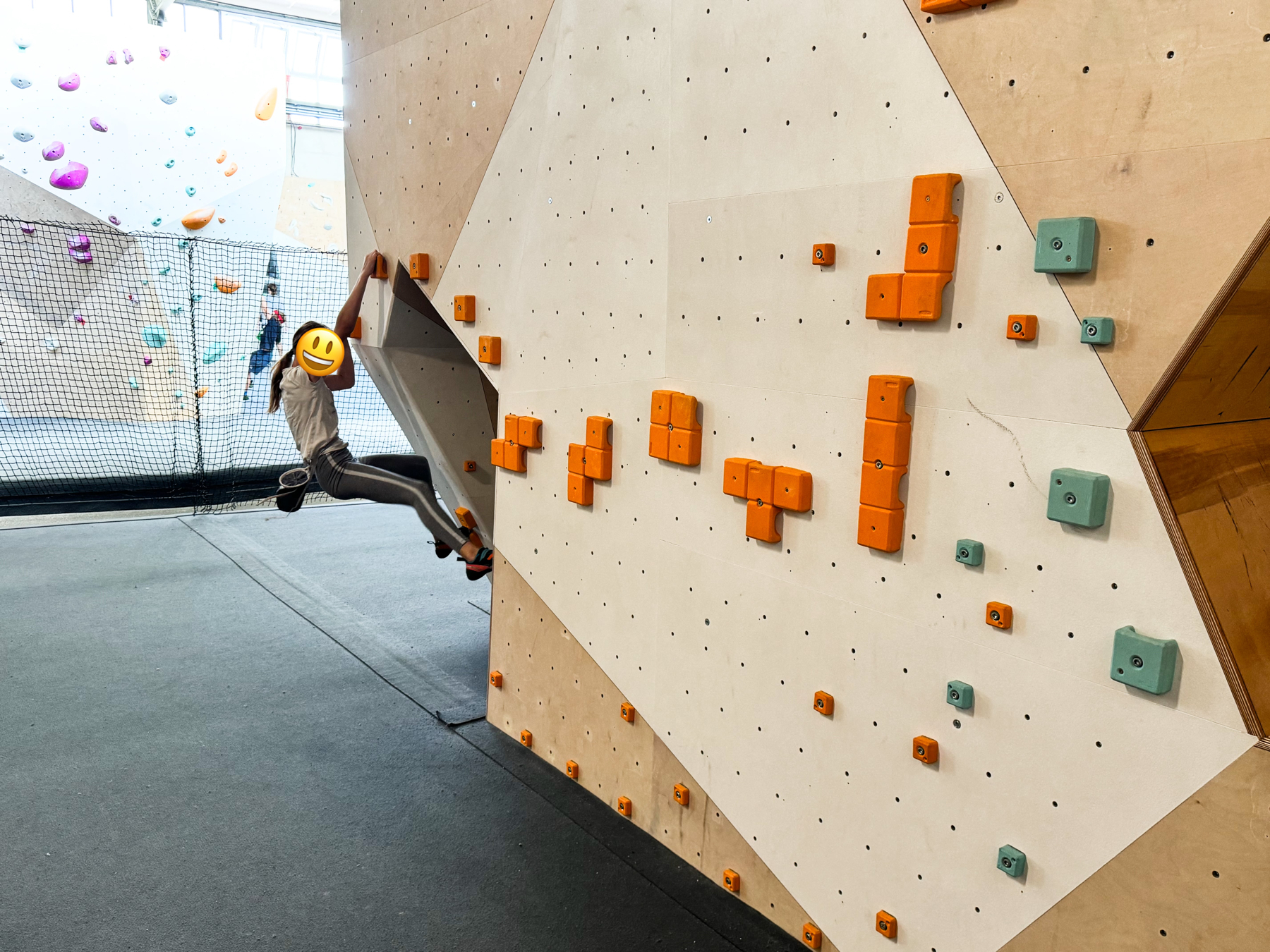 A person wearing climbing shoes is bouldering on an indoor climbing wall with orange and green holds. The climbing facility features padded flooring and additional climbing walls in the background. The climber’s face is covered with a smiley face emoji