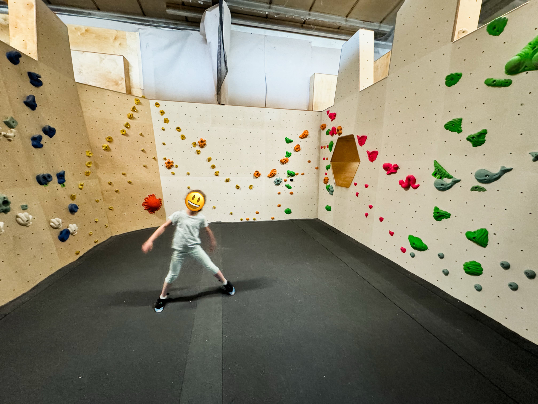 A child standing inside an indoor rock climbing gym, wearing a smiley face emoji over their face. The climbing walls feature various colored holds in blue, yellow, orange, pink, green, and gray. There is a hexagonal wooden structure inset into