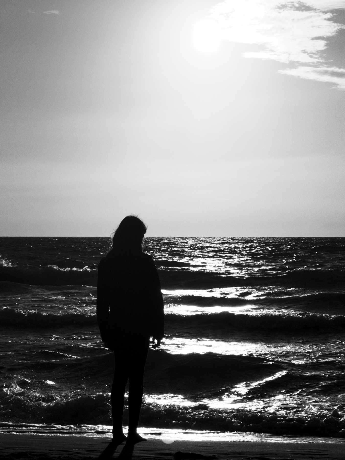 Silhouette of a person standing on a beach, facing the ocean during sunset or sunrise. The sky and water have a bright, reflective quality, and the overall appearance is in black and white.
