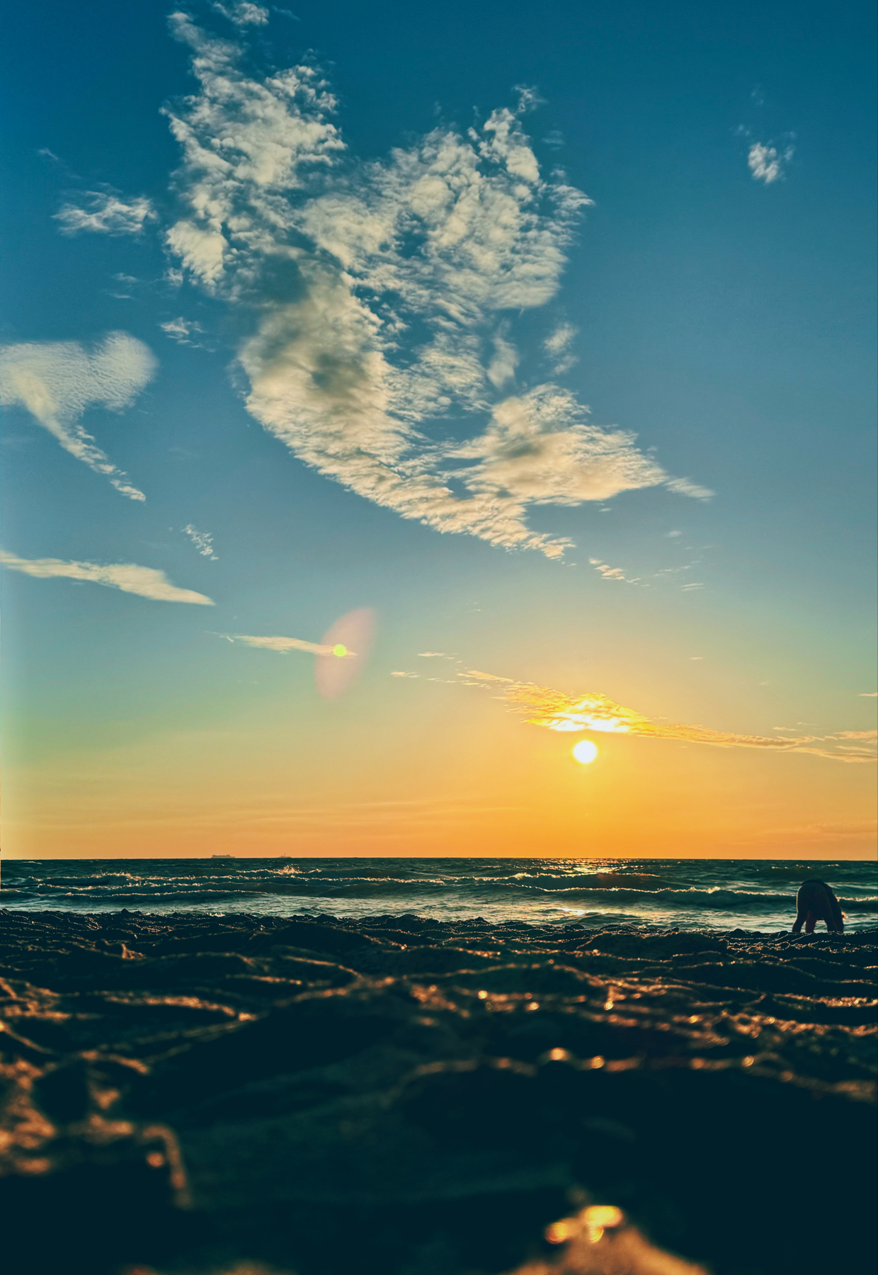 Beach scene at sunset with the sun low on the horizon, a person bending down near the water, and scattered clouds in the sky. Waves gently roll onto the shore, with golden light reflecting off the water and wet sand.