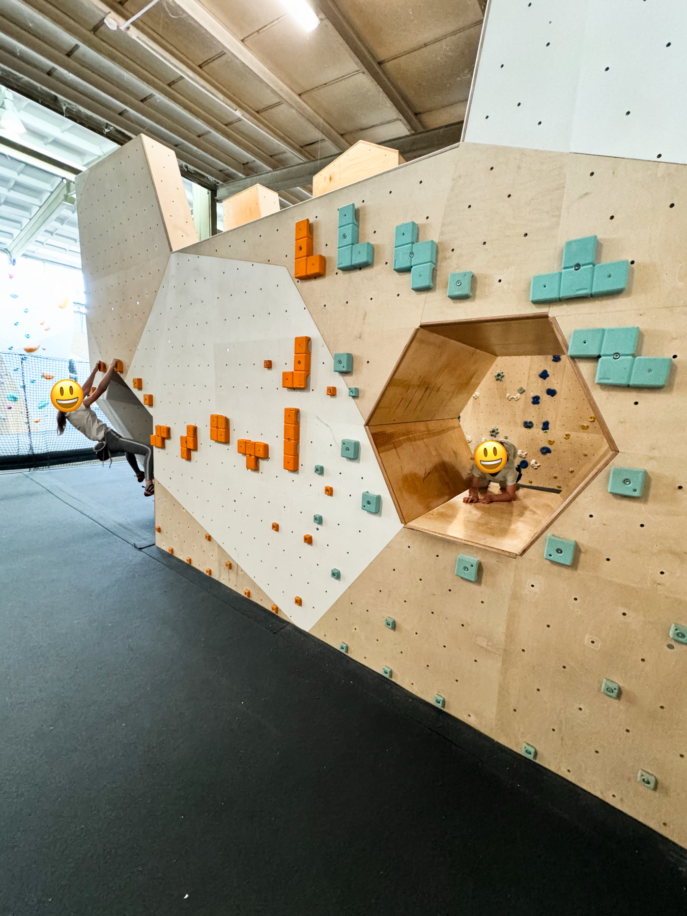 Indoor climbing gym with a wooden climbing wall featuring orange and turquoise holds. A person is climbing on the left side, and another individual is sitting inside a hexagonal cutout on the wall.