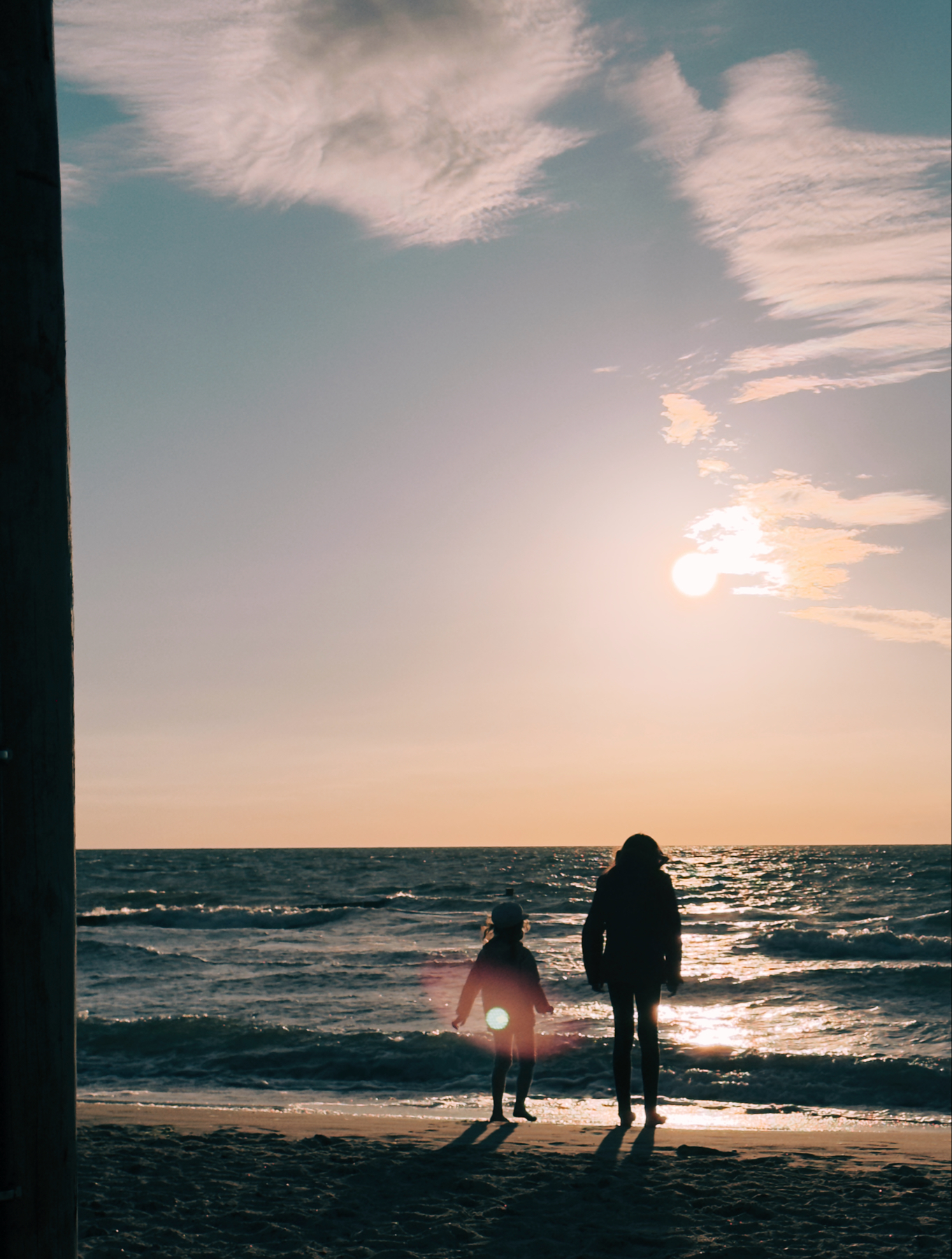 Silhouettes of two people, one of them a child, standing on a beach at sunset, with the ocean and sun in the background. The sky is partly cloudy, and their shadows stretch across the sand.