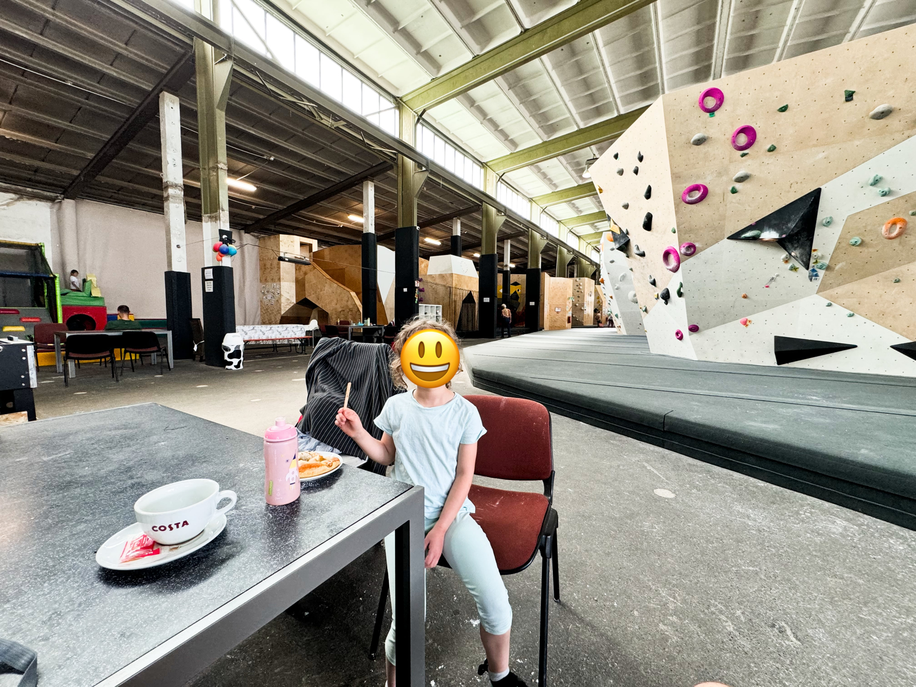 A young child sits at a table in an indoor climbing gym, eating food with a drink bottle and a cup of Costa coffee on the table. The gym features bouldering walls and a children's play area in the background.