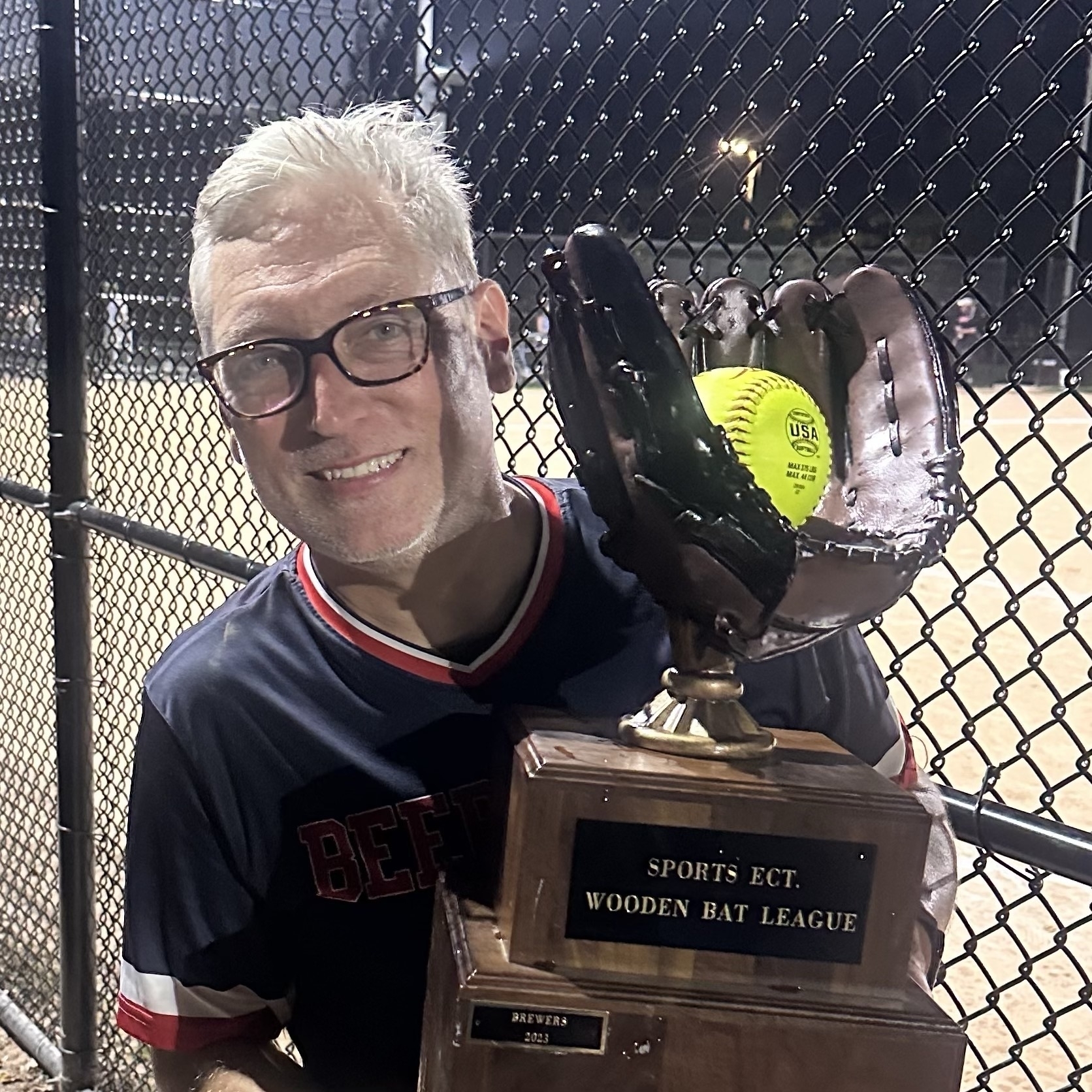 Selfie of a caucasian man with white hair, glasses, smiling while holding a trophy in the shape of a softball mitt with a yellow ball in it.