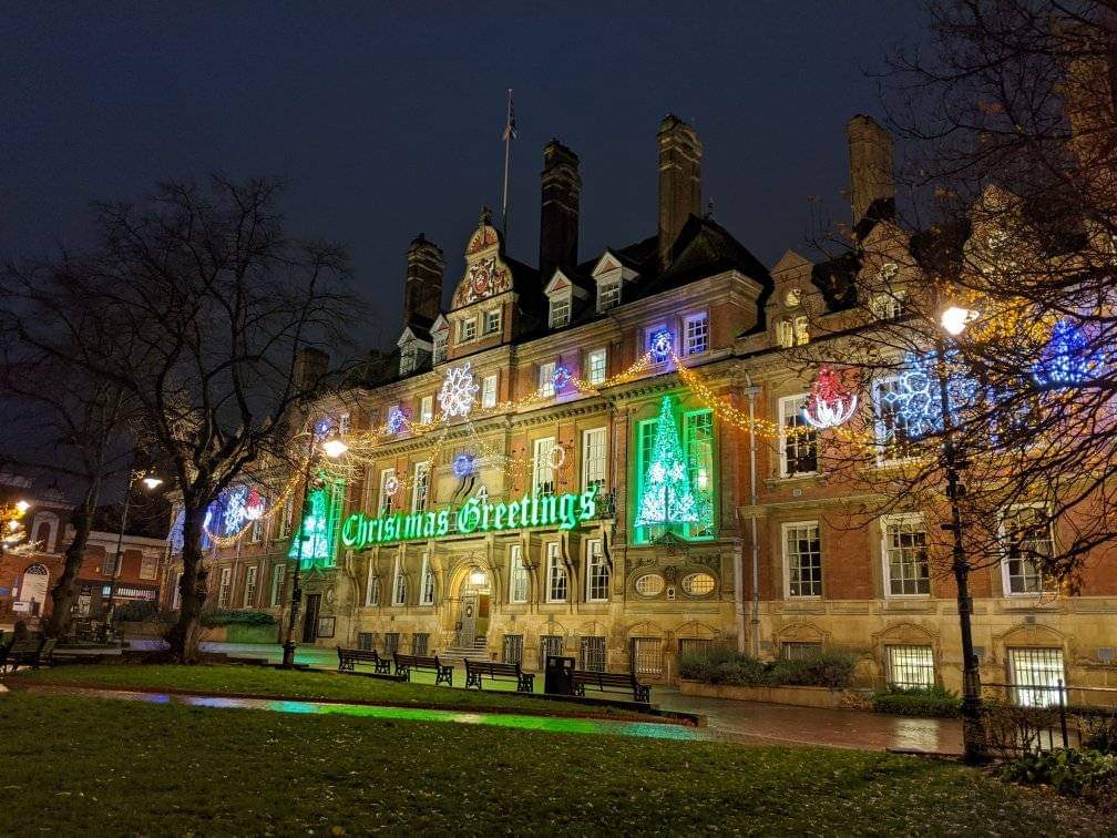 A night-time shot of Leicester Town Hall decorated with Christmas lights and a huge lit-up green neon Chrismas Greetings sign across the facade.