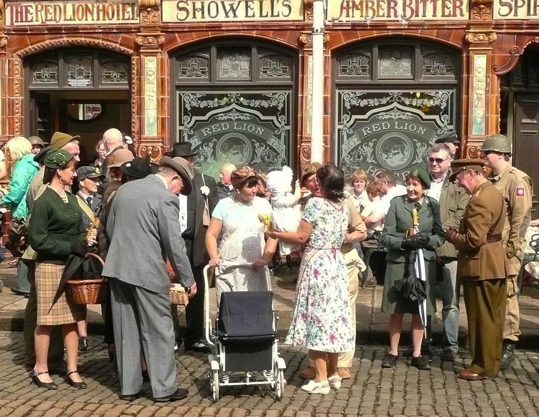 A group of people all dressing in 1940s clothing are chatting and socialising outside the Red Lion, a vintage-looking public house. 