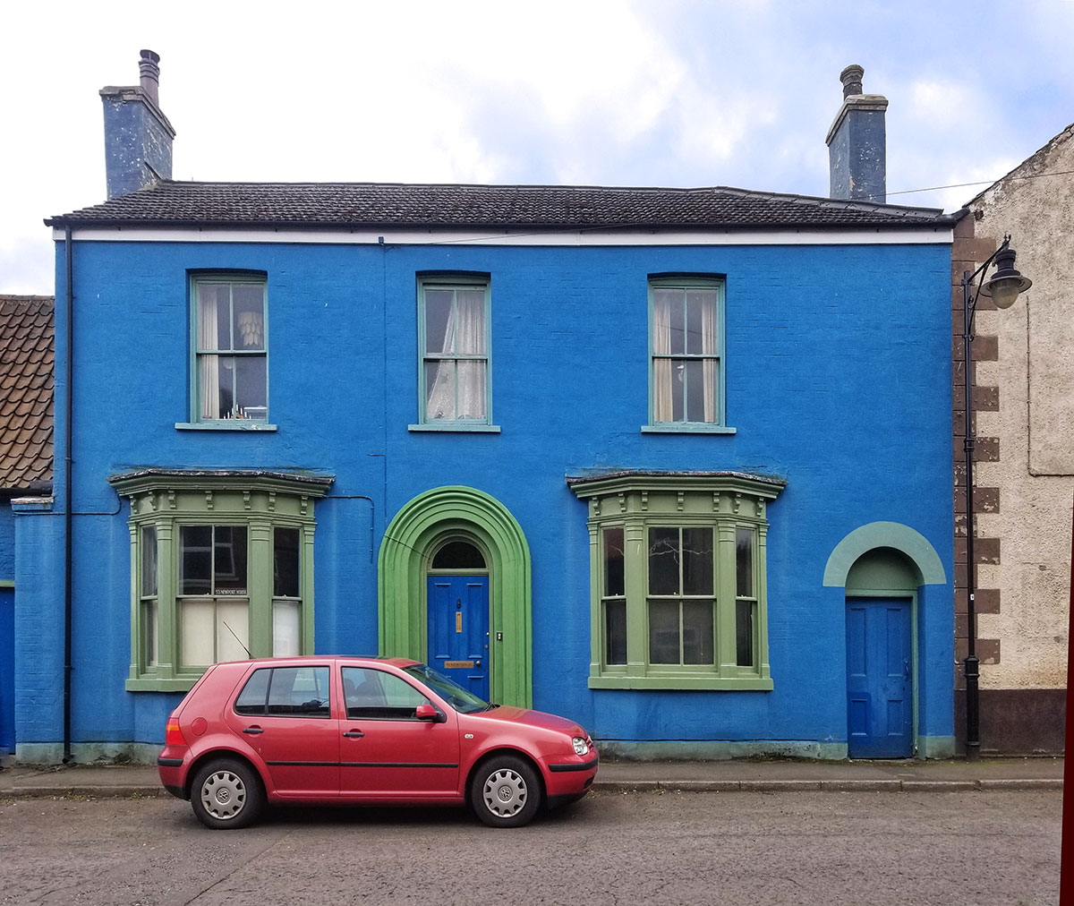 A red park is parked on the road outside a bright blue house with green painted window frames.