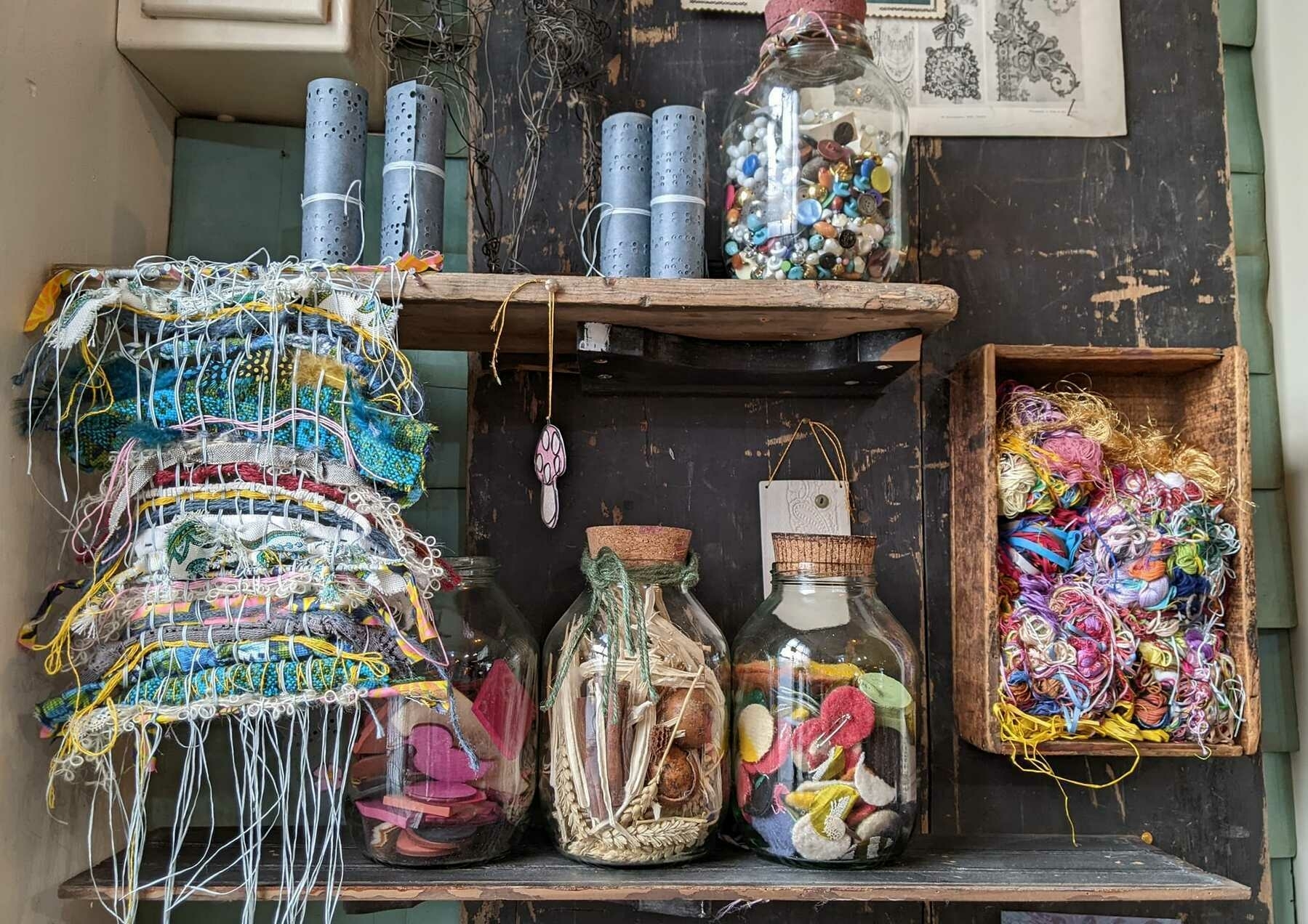 A pile of fabric scraps with lose hanging threads, a shelf holding jars of buttons and felt decorations for sewing