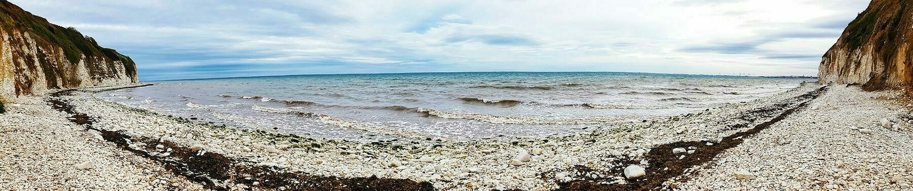 Panoramic view looking out to sea from a beach covered in white stones and pebbles. Either side are chalk cliffs.