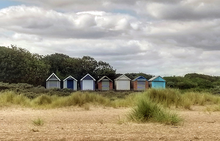A row of beach huts along and empty beach, with marram grass in the foregroud and a dark, threatening sky