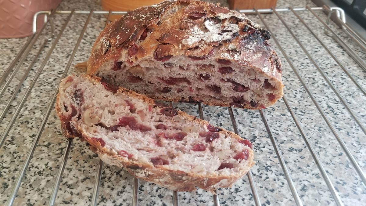 A round rustic cranberry and walnut loaf that has a slice cut from it, showing the fruit-studded inside.