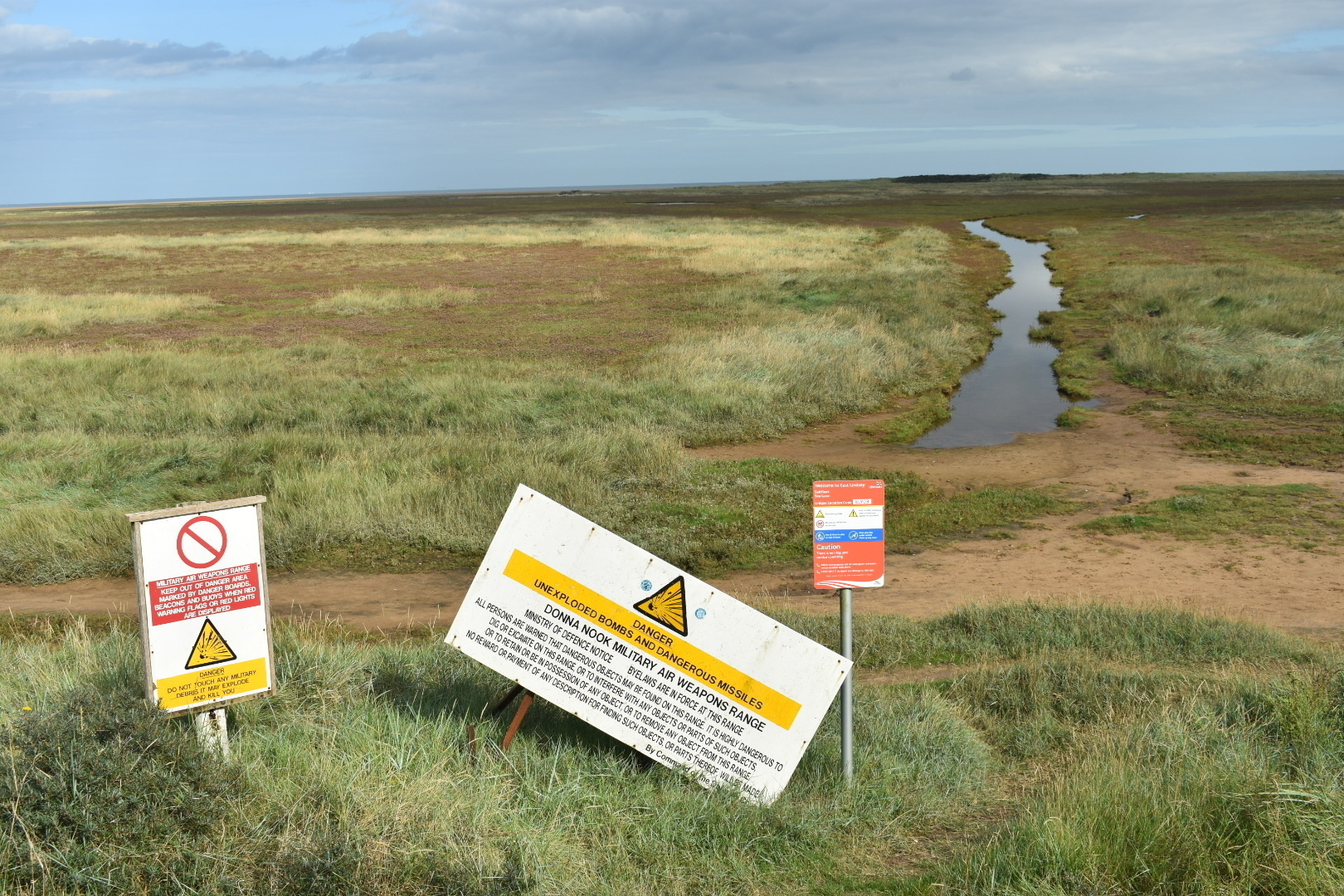 A panoramic view of mudflats and salt marsh with a creek winding into the distance. In the foreground are two warning signs that the area is a military air weapons range and there may be unexploded bombs!