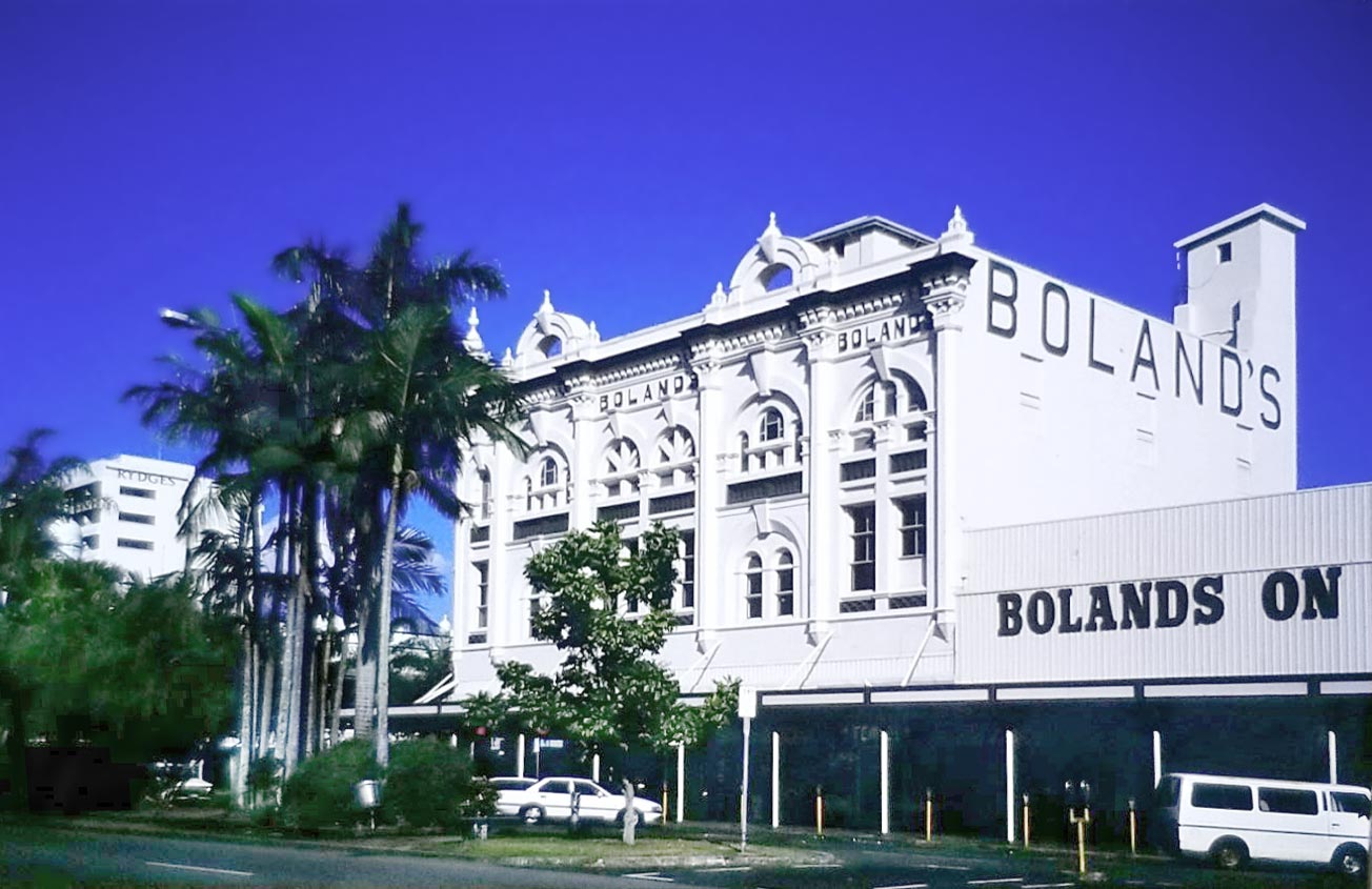 Bolands Center in Cairns, Queensland, Australia. An ornate, grand white building against a bright blue sky, with a row of palm trees in front.