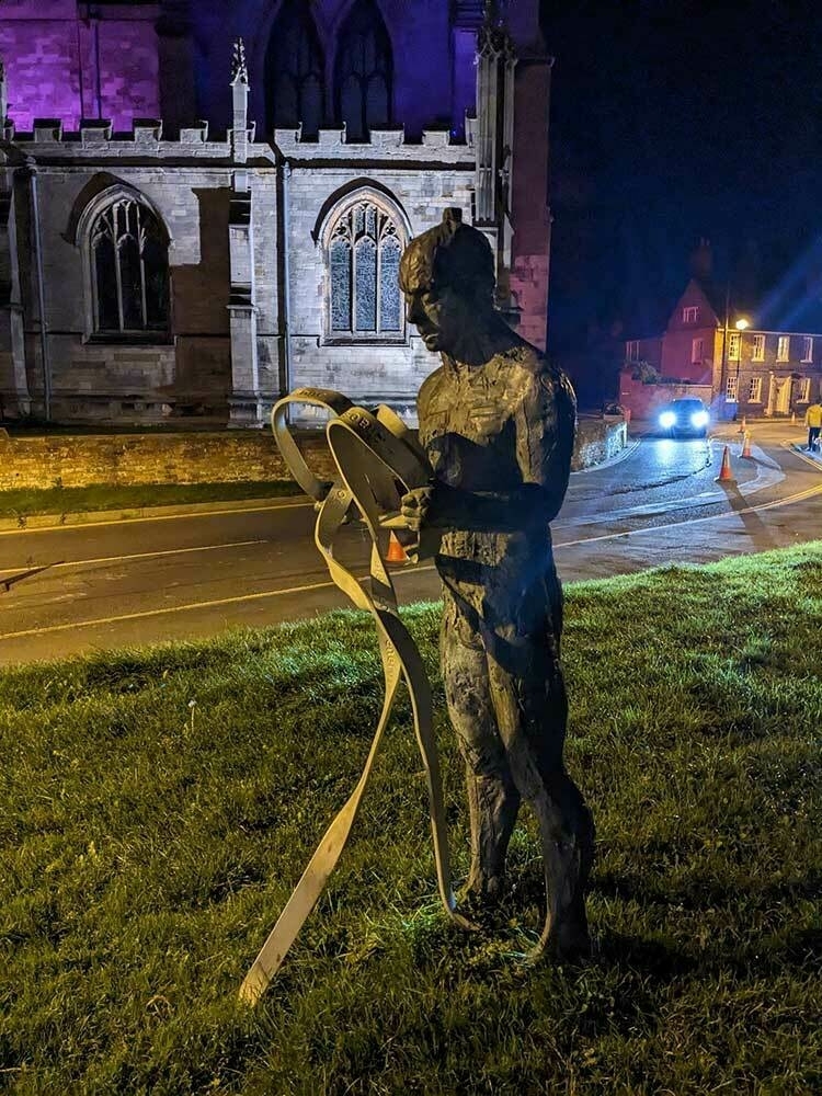 A nighttime scene of one of the Meridian Line sculptures in Louth, in front of a Church which is illuminated bu coloured spotlights.