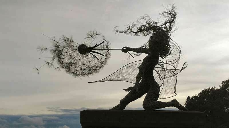 A wire sculpture of a fairy and dandelion silhouetted against a sepia monochrome sky