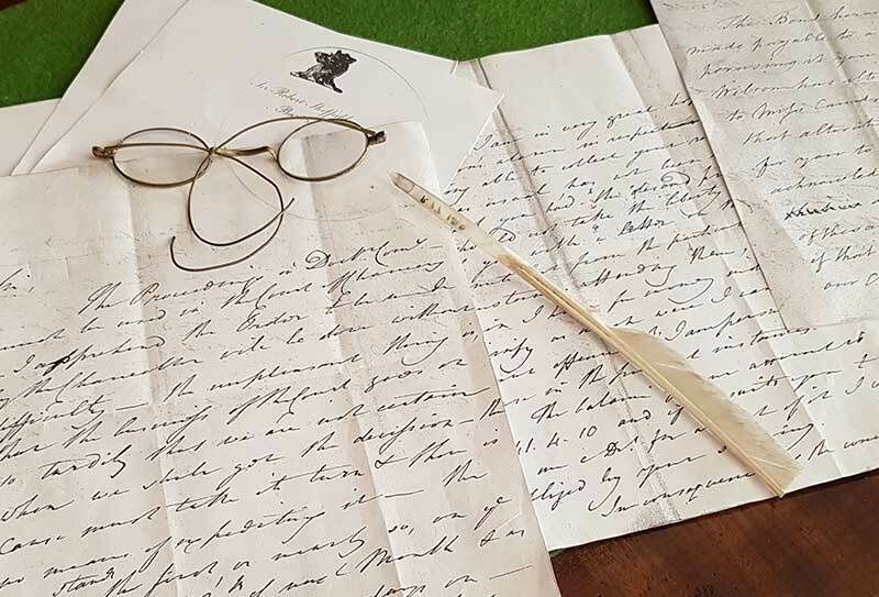 An old letter, handwritten in a beautiful script with a pair of wire spectacles and a quill pen resting on it