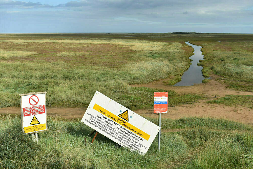 A view looking out over the saltmarshes near the Donna Nook nature reserve. In the foreground is an MOD board warning to look out for unexploded bombs.
