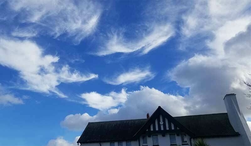 A bright blue sky filled with wispy white clouds, seen above the roof of a house