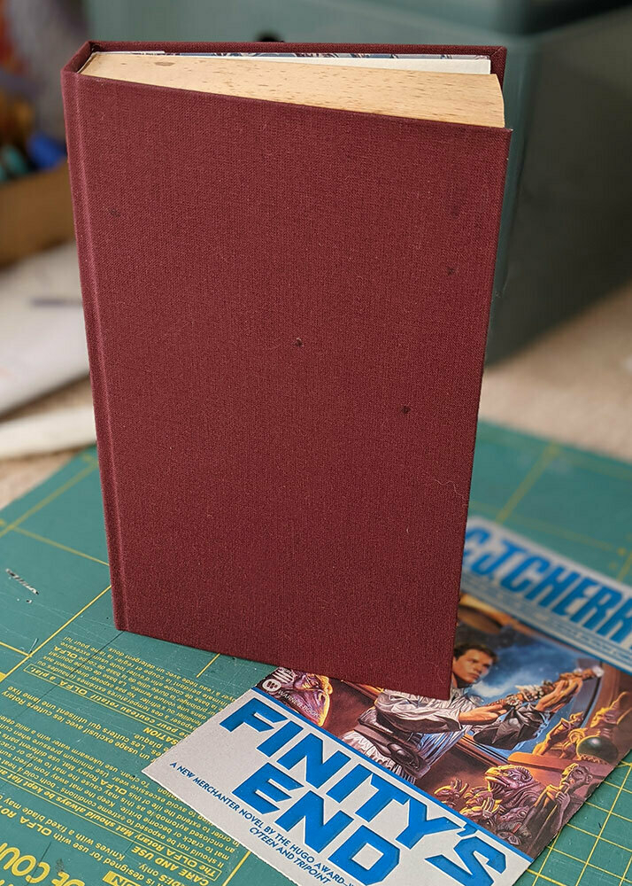 A copy of a CJ Cherryh novel that has been hand-bound by myself in red bookcloth, lying on the original printed cover which was removed.