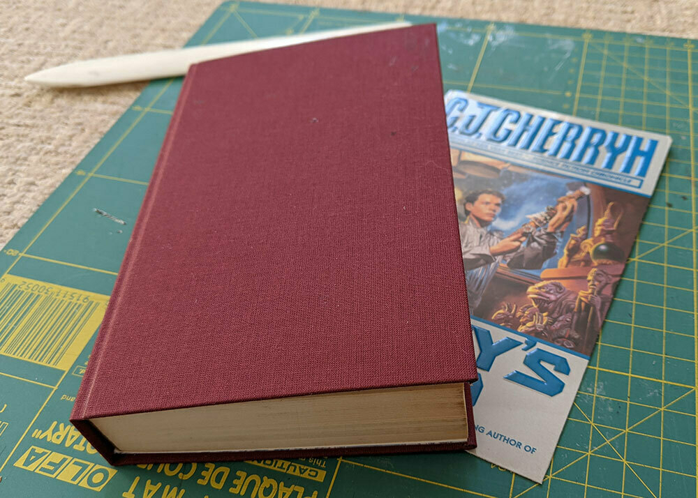 A copy of a CJ Cherryh novel that has been hand-bound by myself in red bookcloth, lying on the original printed cover which was removed.