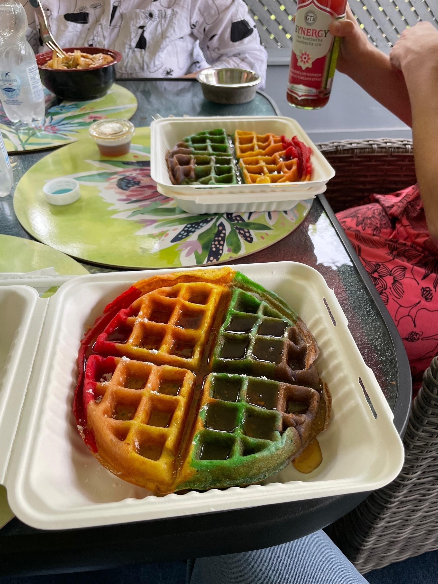 Two big waffles in take our containers that are coloured in a rainbow of red, yellow, green, and purple like a pride flag. Drizzled with delicious maple syrup.