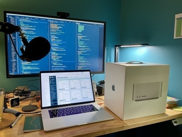 A messy desktop with a brand new Mac Studio box beside a 2009 Mac Book Pro and a 32 inch display showing code in the background.