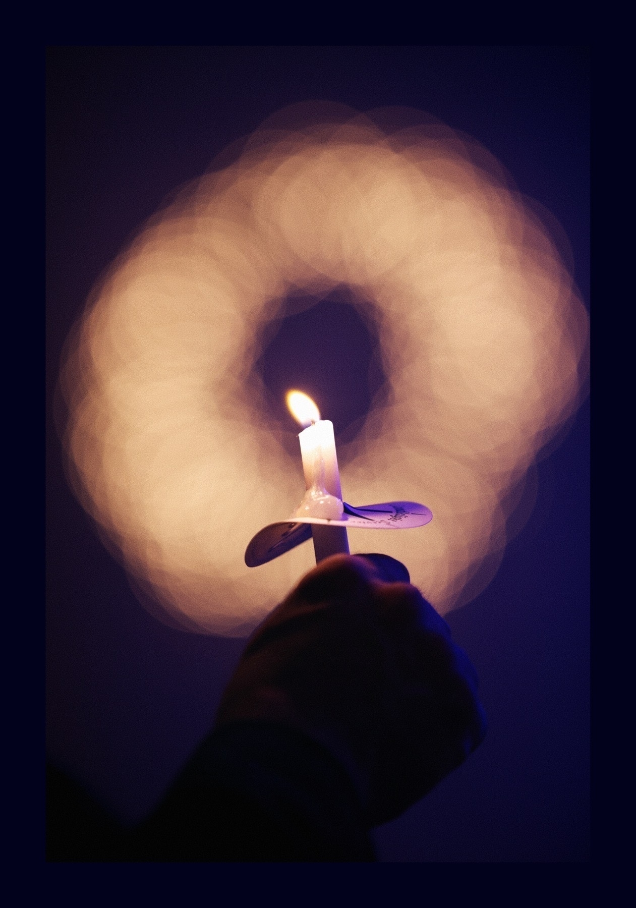 A lit candle is held in front of an out-of-focus ring of Christmas lights.