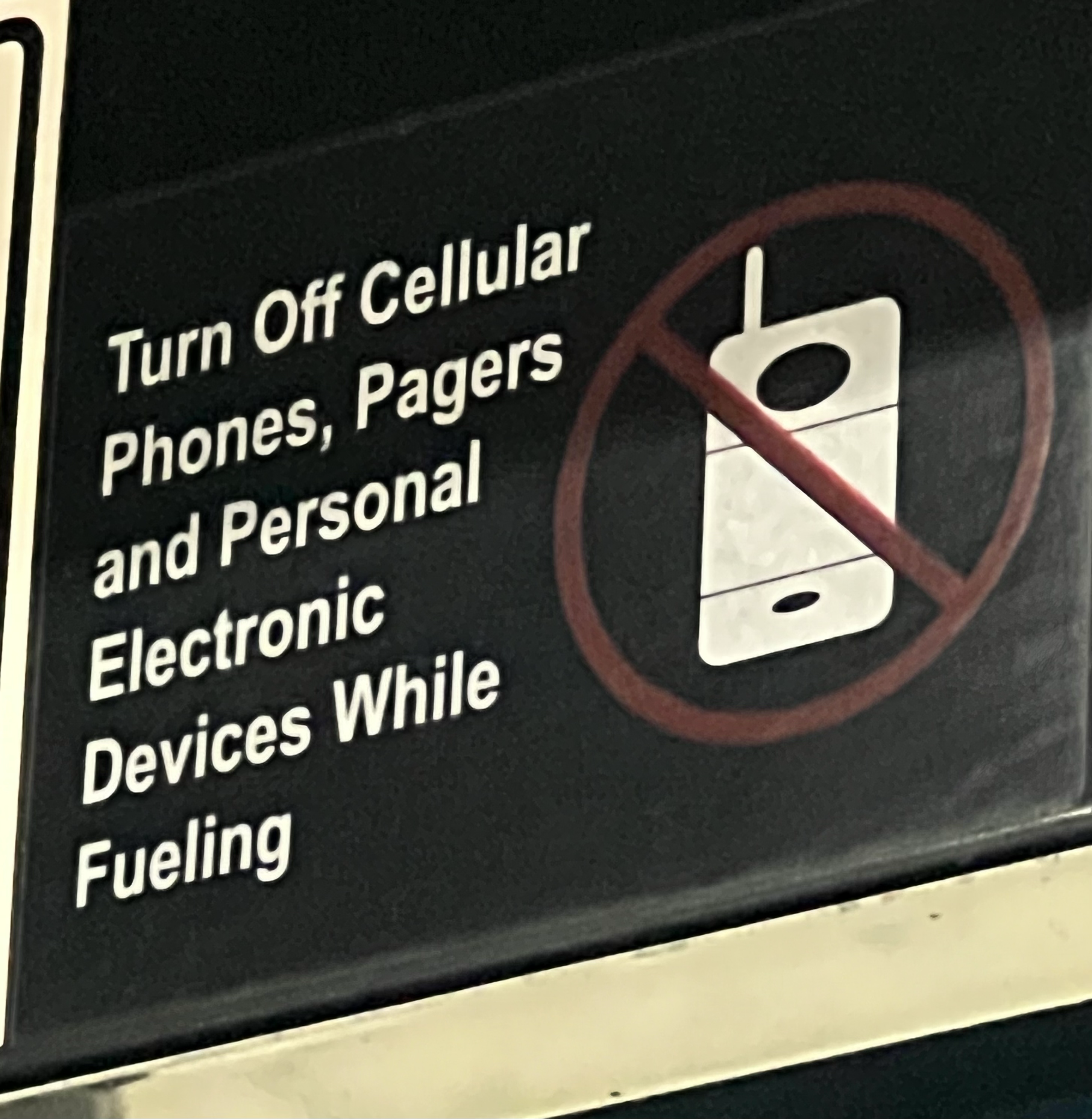 Gas station sign that says “Turn off cellular phones, pagers, and personal electronic devices while fueling.” Next to it is an image of an old cell phone with a line through it.