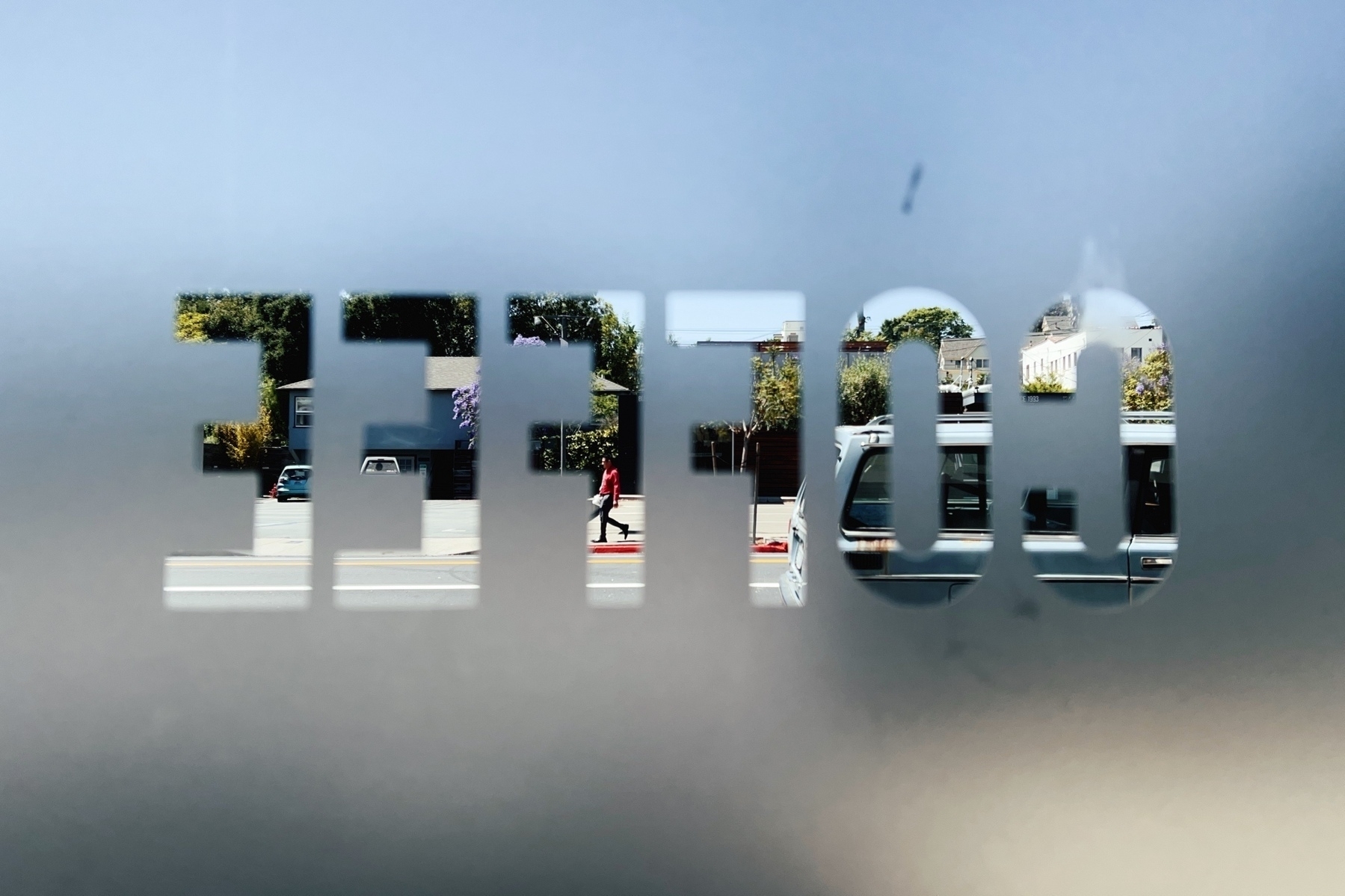 Photo taken through a coffee shop window. The window reads “COFFEE” backwards. In the distance someone walks down the sidewalk in a red sweater. A car drives by.