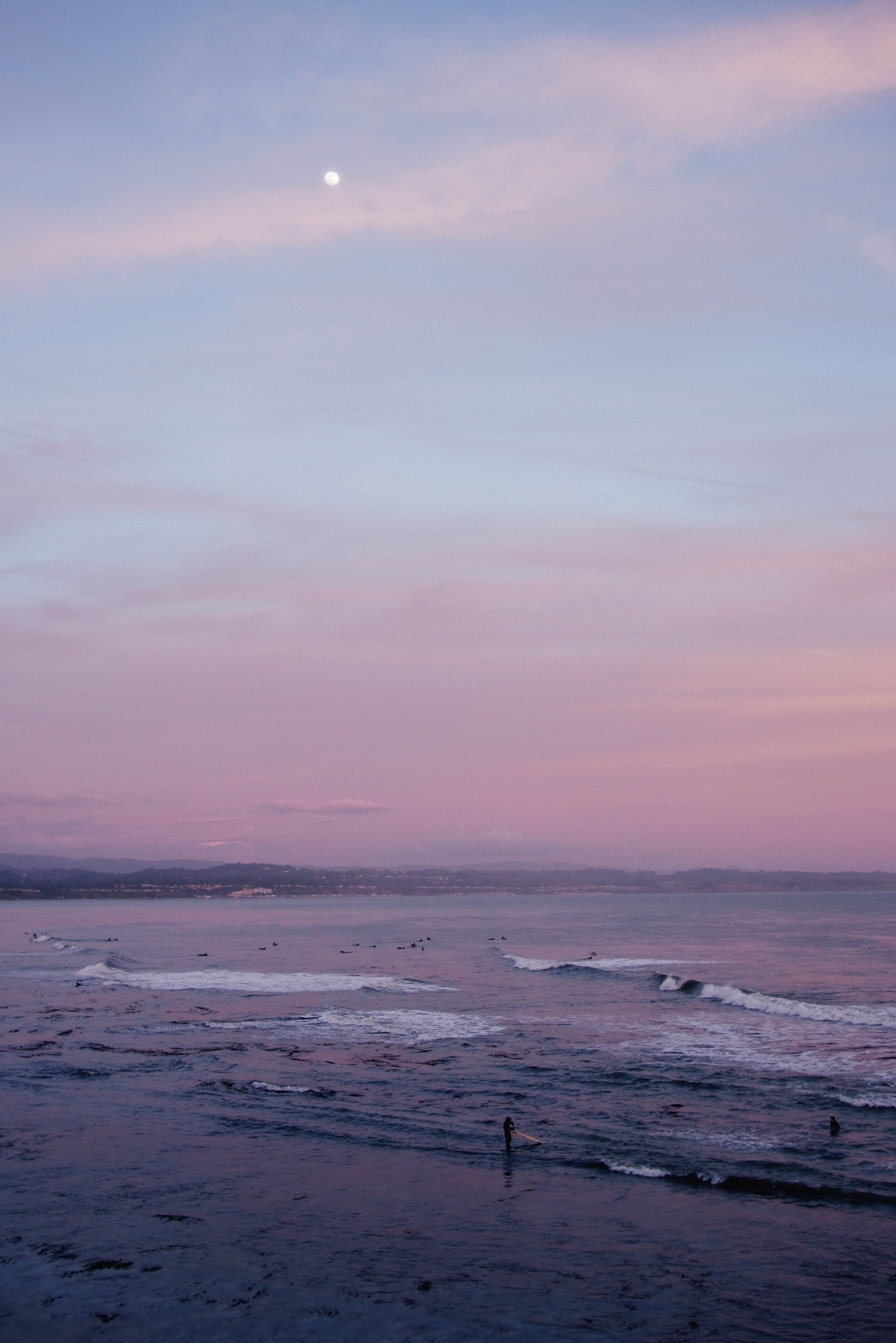 Sunset. A surfer pulls his board out of the waves. The sky is pink and so is the sand reflecting it. The moon hangs as a small dot above it all.