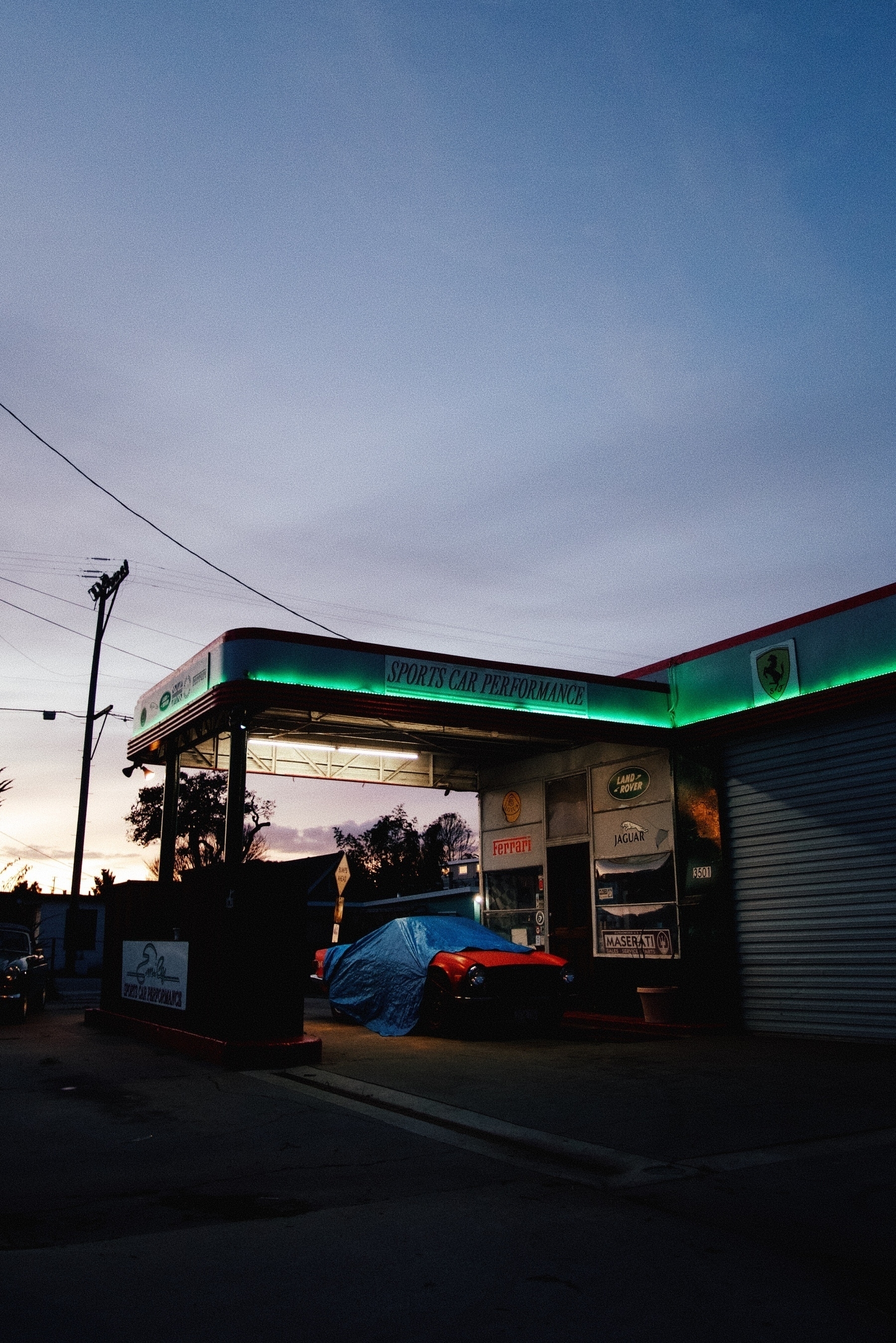 An auto shop just after sunset. A sign reads Sports Car Performance. Green trim lighting illuminates this sign and others. A red car is halfway covered underneath it.