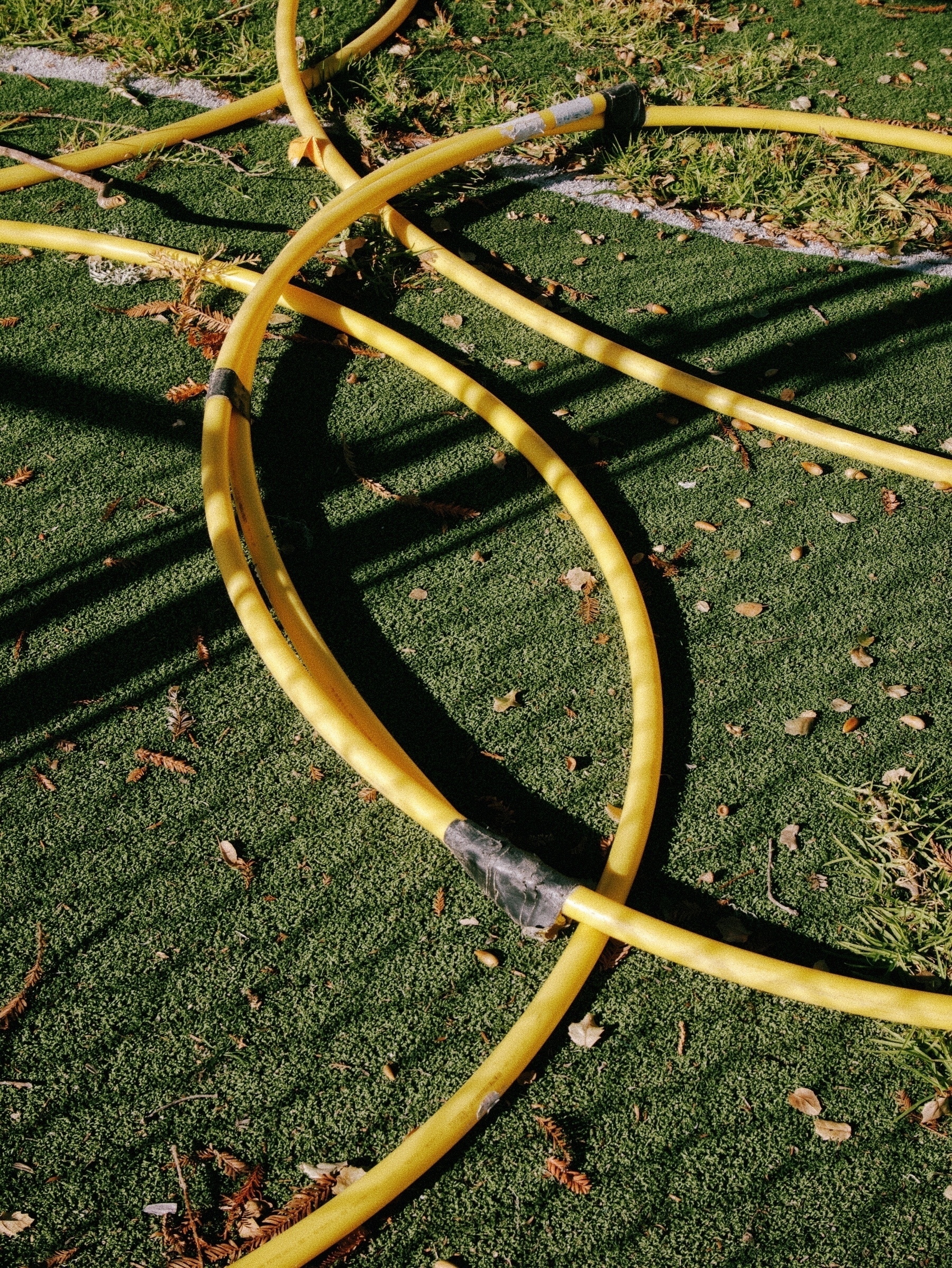 Color photograph of broken yellow hula hoops lying on fake grass. A shadow of a chain link fence covers them.