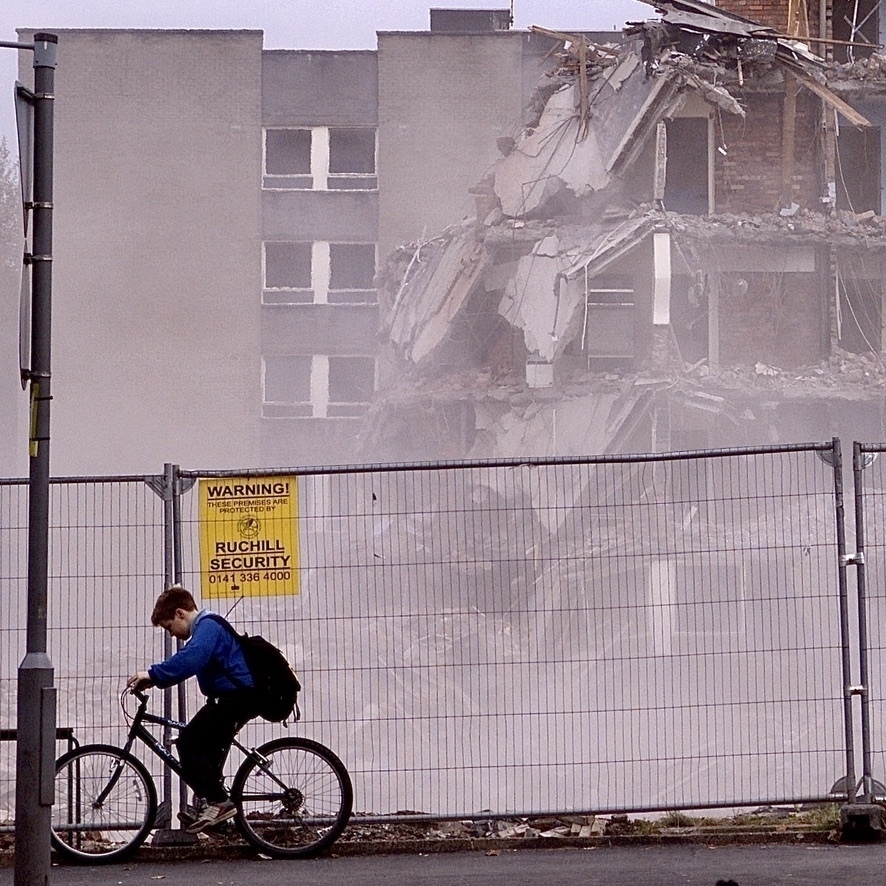 A photograph. In the background is a half-destroyed block of flats. In the foreground is a security fence, and to the left in front of that is a boy on a bike looking downwards